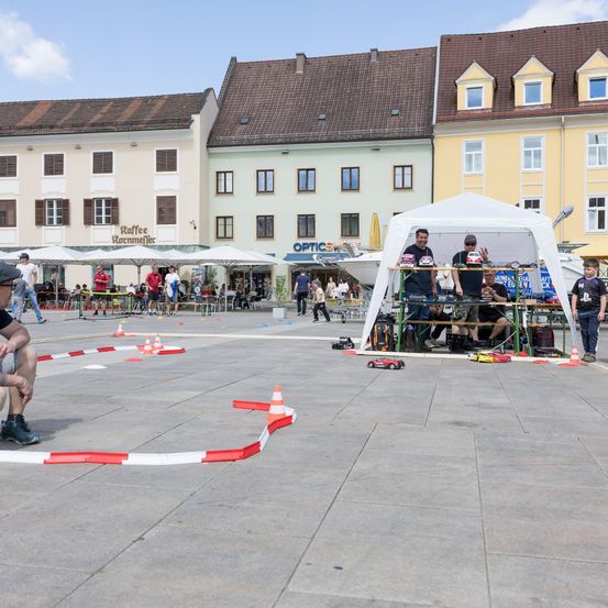 Ein Autorennen auf einer Rennstrecke in einem Stadtplatz, das von Menschen beobachtet wird. Ein weißes Zelt hat mehrere Personen im Inneren. Im Hintergrund sind Gebäude und Cafés.