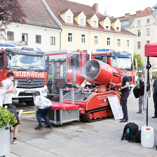 Mehrere Menschen interagieren mit einer großen roten Maschine vor Feuerwehrautos, während andere in der Nähe stehen. Die Maschine hat Text und einen großen Lüfter, und es gibt Gebäude und Bäume im Hintergrund.