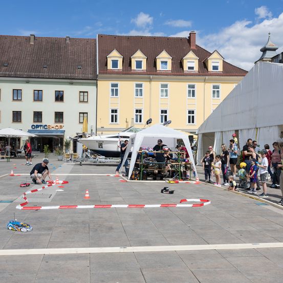Eine Gruppe von Menschen versammelt sich auf einem Stadtplatz für ein Rennen mit ferngesteuerten Autos. In der Nähe befinden sich mehrere Zelte und ein Boot.