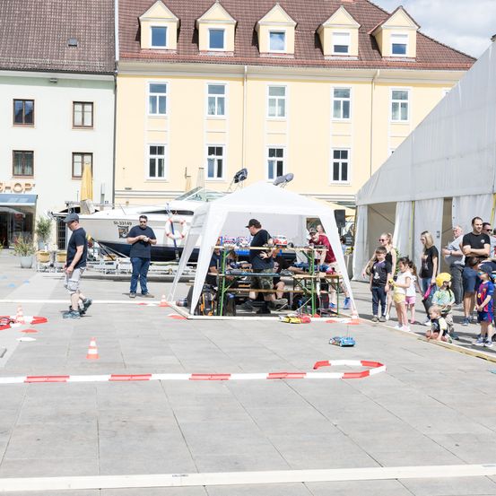 Eine Menschenmenge versammelt sich um ein weißes Zelt auf einem Stadtplatz. Die Leute beobachten ein Rennen mit kleinen Autos auf einer provisorischen Rennstrecke. Im Hintergrund befinden sich Gebäude mit vielen Fenstern.