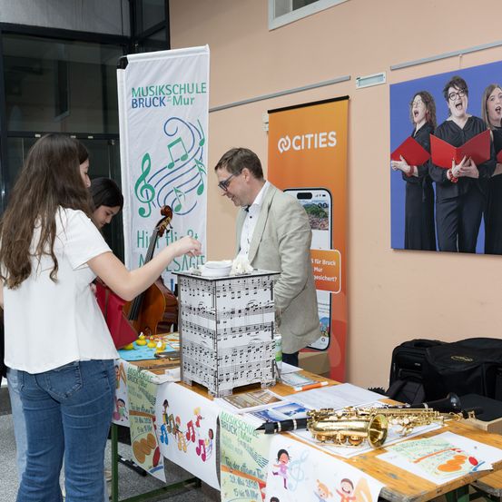 Ein Infostand einer Musikschule mit Cello, Saxophon und verschiedenen Plakaten steht in einem Raum mit Glaswänden.
