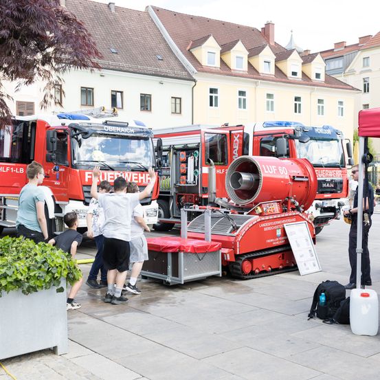 Eine Gruppe von Menschen, darunter Kinder, steht vor Feuerwehrautos, wobei eine Person die Hände hebt. Ein Feuerwehrmann steht neben einer roten Maschine. Ein weißer Beutel und ein weißer Plastikbehälter liegen auf dem Boden.