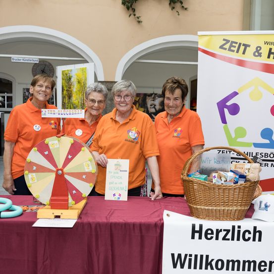 Vier Frauen in orangefarbenen T-Shirts stehen hinter einem Tisch mit einem Drehrad, einem Schild mit der Aufschrift 'Zeit & Wir' und einem Korb. Der Tisch ist mit einem roten Tuch bedeckt.