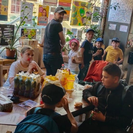 Mehrere Kinder und Erwachsene in einem Café mit einem Mann, der hinter ihnen steht, essen und trinken. Topfpflanzen und Poster im Hintergrund.