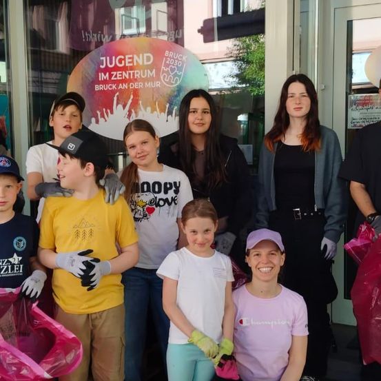 Mehrere Personen stehen vor einem Gebäude und halten Plastiktüten. Zwei Frauen stehen vorne und lächeln für ein Foto. Ein Schild steht für Jugend im Zentrum Bruck an der Mur.