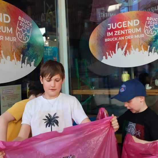 Zwei Jungen halten rosa Tüten in der Hand, eine mit einem Palmenbaumdesign, vor einem Geschäft mit farbenfrohen Schildern. Hinter ihnen ist teilweise eine Person in einem gelben T-Shirt zu sehen.