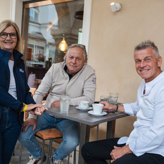 Drei Erwachsene sitzen auf Stühlen an einem Tisch vor einem Café und posieren für ein Foto. Auf dem Tisch stehen Tassen und Gläser.
