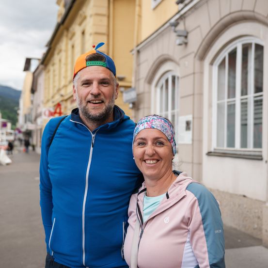 Ein Mann und eine Frau stehen gemeinsam auf einem Bürgersteig, beide in Sportkleidung und lächeln für ein Foto. Der Mann hat einen bunten Hut und einen Bart. Hinter ihnen steht ein Gebäude mit Fenstern und einem Schild.