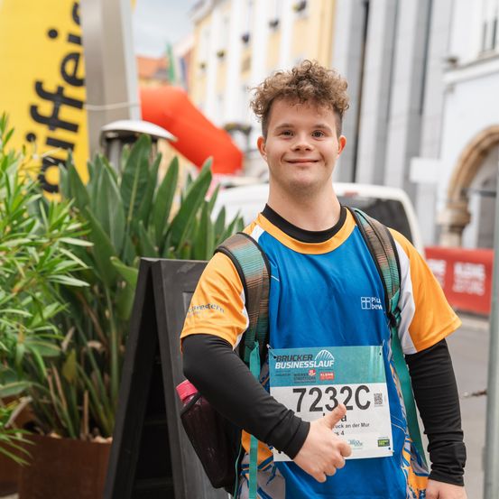 Ein lächelnder Mann in einem blauen Shirt mit der Nummer 7232C steht draußen mit einem Rucksack und einer Pflanze im Hintergrund.