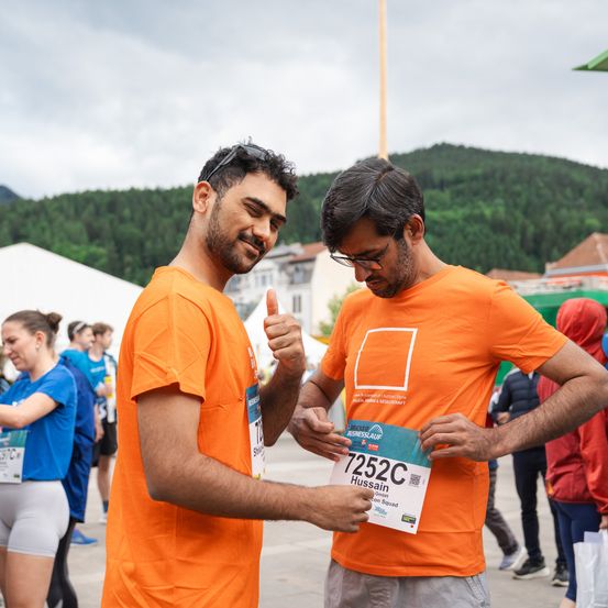 Zwei Männer in orangefarbenen T-Shirts mit der Nummer 7252C, wahrscheinlich bei einem Marathon, geben Daumen hoch. Im Hintergrund laufen Leute mit Zelten, einem Berg und Gebäuden vorbei.