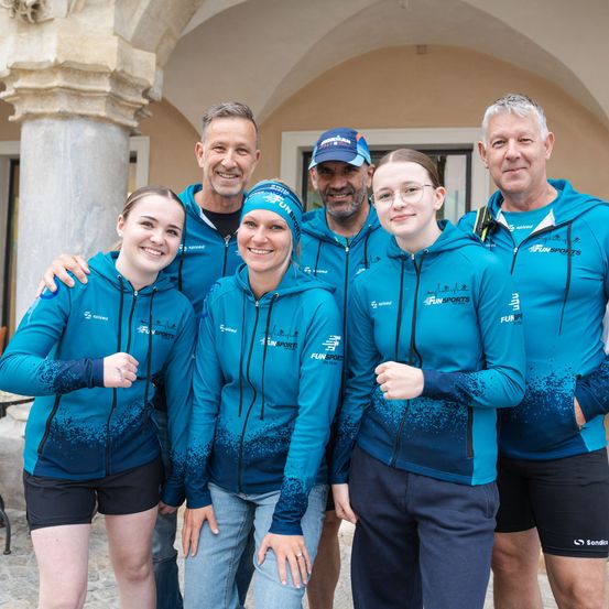 Eine Gruppe von sieben Personen, einschließlich Männer und Frauen, steht lächelnd vor einem Gebäude und trägt passende blaue Sportjacken.