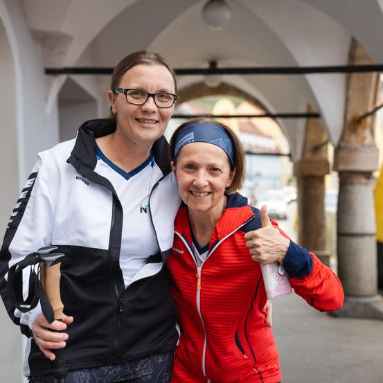 Zwei Frauen stehen zusammen draußen, lächeln und posieren für ein Foto. Eine trägt eine schwarz-weiße Jacke und hält einen Trekkingstock. Die andere in einer roten Jacke gibt einen Daumen hoch.