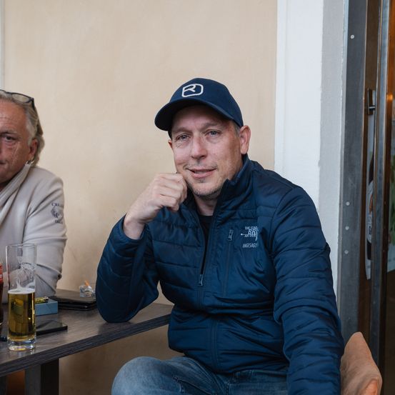 Ein Mann mit einer blauen Kappe und einem Jackett sitzt an einem Tisch mit einem Glas Bier, während ein anderer Mann mit einer Brille auf dem Kopf neben ihm sitzt.