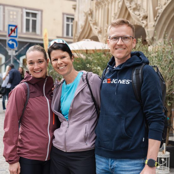 Drei Personen auf einem Stadtplatz. Zwei Frauen und ein Mann. Sie lächeln in die Kamera. Der Mann trägt eine Brille und einen Rucksack.