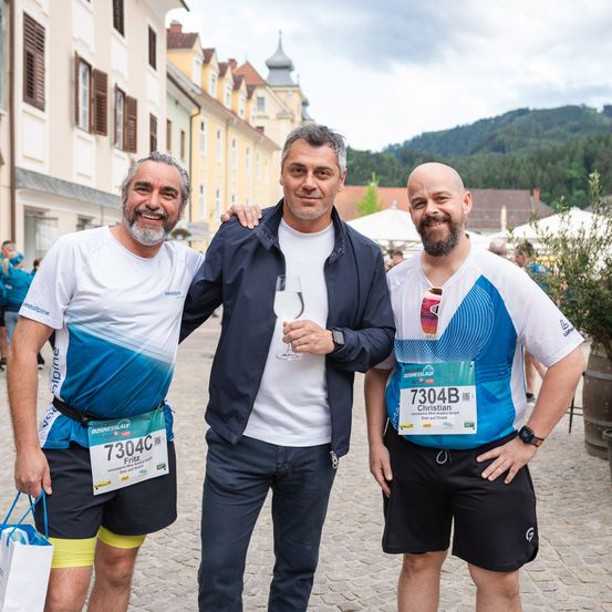 Drei Männer stehen lächelnd zusammen auf einer Kopfsteinpflasterstraße. Der Mann in der Mitte hält ein Weinglas. Alle drei tragen Laufwesten mit Startnummern. Im unscharfen Hintergrund sind Gebäude und Berge zu sehen.