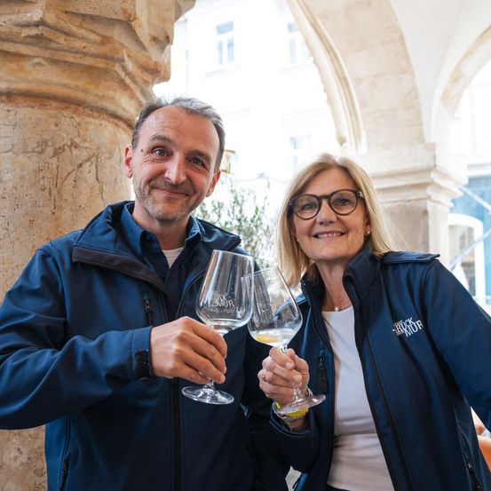 Ein Mann und eine Frau, beide in blauen Jacken, stehen nebeneinander und halten Weingläser. Sie lächeln in die Kamera, wahrscheinlich bei einer Weinveranstaltung.