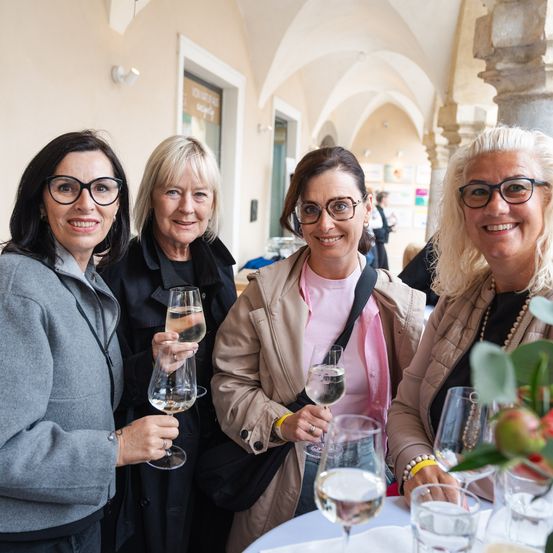 Vier Frauen mit Brillen lächeln und halten Weingläser an einem Tisch mit Blumenarrangement. Sie stehen in der Nähe eines gewölbten Durchgangs und einer Wand mit einem Schild.