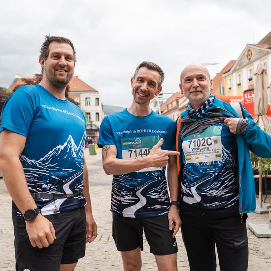 Drei Männer stehen auf einer Kopfsteinpflasterstraße, alle tragen Laufshirts. Der Mann in der Mitte zeigt mit seinem Finger. Dahinter ist ein unscharfer Hintergrund von Gebäuden und Grün.