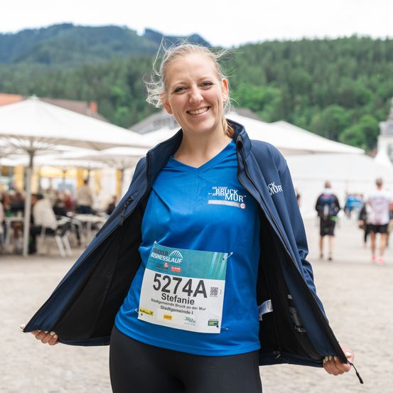 Eine Frau steht lächelnd im Freien, trägt ein blaues T-Shirt mit der Nummer 5274A. Sie hat eine offene blaue Jacke an und hält die Jacke mit beiden Händen. Im Hintergrund sind Personen und Zelte zu sehen.