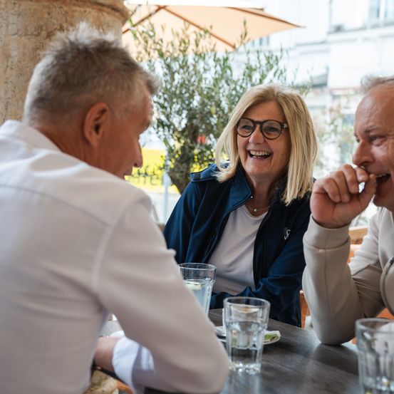 Drei Personen sitzen an einem Tisch im Freien, wahrscheinlich in einem Restaurant. Eine Frau trägt eine Brille und lächelt. Zwei Männer schauen sie an, einer hält sein Kinn. Der Tisch hat Wassergläser. Dahinter steht ein Baum und ein Gebäude.