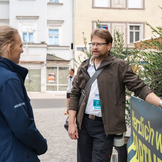 Ein Mann und eine Frau unterhalten sich im Freien. Er trägt eine Brille und eine braune Jacke. Hinter ihnen ist ein Gebäude zu sehen.