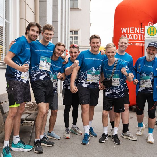 Eine Gruppe lächelnder Athleten in passenden blauen Shirts mit Laufnummern posiert für ein Foto im Freien. Sie stehen auf einem Bürgersteig vor einem großen roten Schild.
