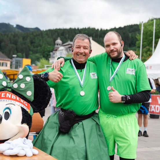 Zwei Männer in grünen Outfits und Medaillen posieren für ein Foto mit einer Minnie-Mouse-Attrappe. Sie stehen an einem Ort im Freien, wahrscheinlich bei einer Veranstaltung.