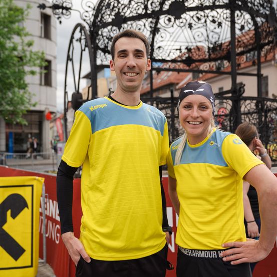 Ein Mann und eine Frau in gelben und blauen Sporttrikots stehen lächelnd nebeneinander. Sie stehen vor einer dekorativen Metallstruktur mit einer Kulisse von Gebäuden.