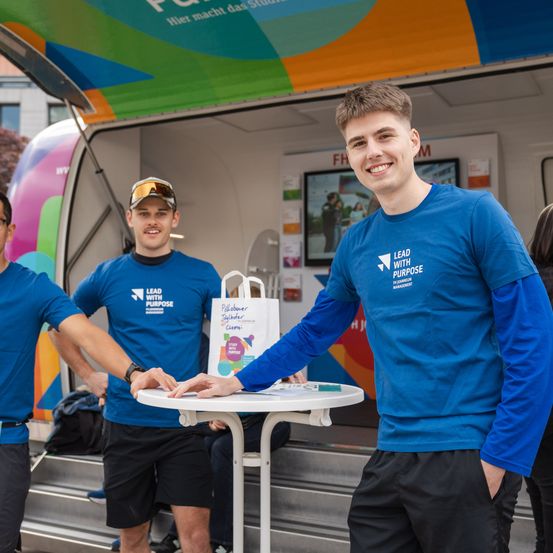 Drei Männer in blauen Shirts stehen vor einem farbenfrohen Imbisswagen. Sie lächeln und halten eine weiße Tasche. Der Mann in der Mitte schüttelt einem anderen Mann die Hand. Hinter ihnen ist ein Monitor an der Wand befestigt.