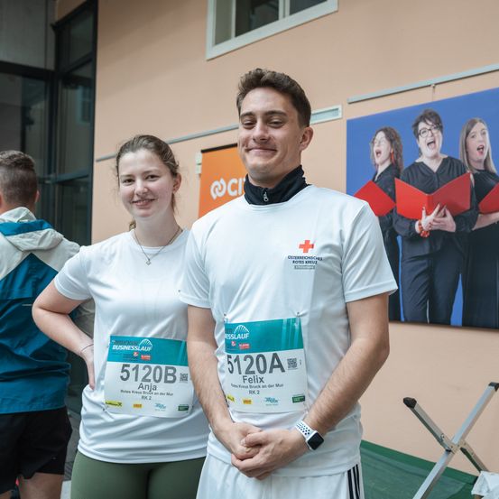 Ein lächelnder Mann und eine Frau stehen in der Nähe einer Wand mit einem Poster singender Frauen. Der Mann trägt ein weißes Hemd mit 'Rotes Kreuz' und Felix 5120A. Die Frau hat einen Startnummern-Aufkleber mit Anja 5120B. Sie sind wahrscheinlich bei einer Marathon-Veranstaltung.