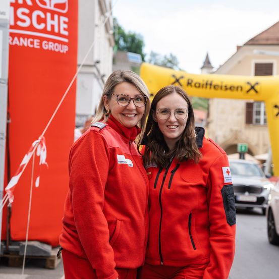 Zwei Frauen in roten Uniformen mit roten Kreuzen auf der rechten Brust stehen zusammen und lächeln. Dahinter ist ein gelber Bogen mit dem Text Raiffeisen. Ein rotes Banner mit dem Text SCHE ist links.