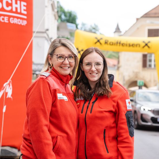 Zwei lächelnde Frauen in roten Jacken stehen nebeneinander auf einer Straße, hinter ihnen ein Auto. Ein roter Banner mit weißem Text und ein gelber Bogen mit dem Wort 'Raiffeisen' sind im Hintergrund.