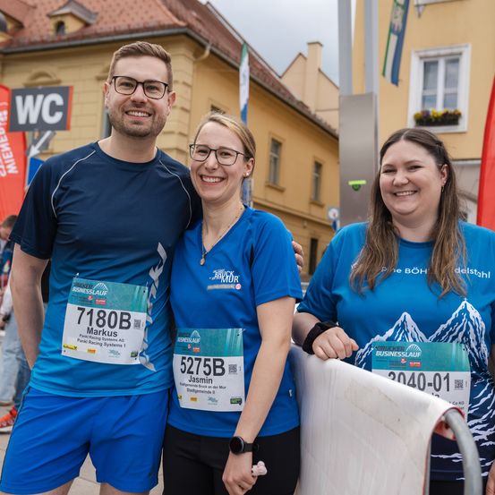 Drei Menschen stehen vor einem Gebäude. Zwei Frauen tragen blaue T-Shirts, eine mit einem Nummern-Bib. Der Mann trägt eine Brille und ein blaues T-Shirt und lächelt. Ein Schild auf Deutsch ist über ihnen.