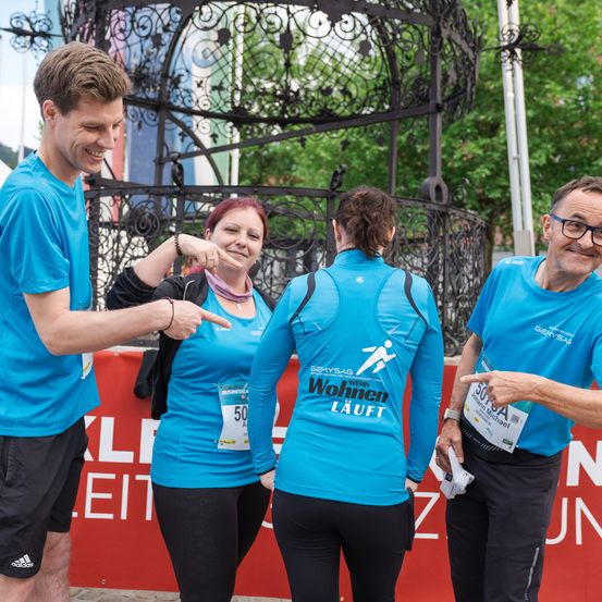 Drei Personen in passenden blauen T-Shirts mit Teilnehmernummern stehen zusammen. Die Frau in der Mitte zeigt auf etwas und lächelt. Hinter ihnen ist ein roter Banner, auf dem Text auf Deutsch steht.