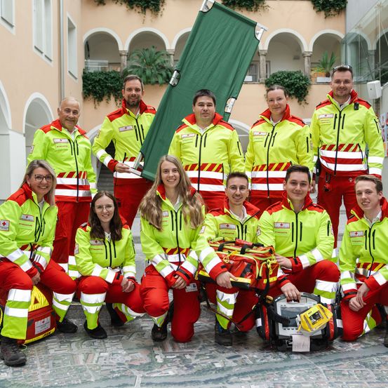 Eine Gruppe von Menschen in gelben und roten Warnwesten posiert für ein Foto im Freien. Sie halten eine Trage und eine medizinische Tasche.