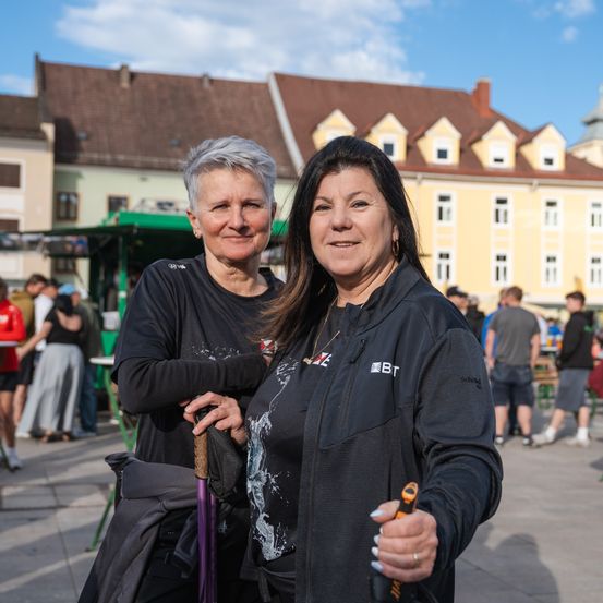 Zwei Frauen stehen auf einem Stadtplatz mit einem großen Gebäude im Hintergrund. Sie lächeln und posieren für ein Foto, eine hält einen Gehstock.