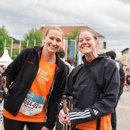 Zwei Frauen mit lächelnden Gesichtern stehen zusammen und halten Skistöcke. Eine trägt eine schwarze Jacke mit orangem Shirt, die andere eine schwarze Kapuzenjacke. Sie stehen vor einem verschwommenen Hintergrund mit Bäumen und einem Gebäude.