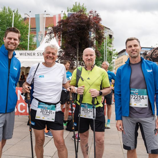 Vier Männer mit Startnummern stehen vor einem Banner. Sie lächeln und scheinen an einem Marathon teilzunehmen. Hinter ihnen sind Menschen und Gebäude.