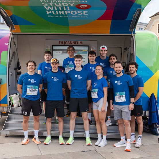 Eine Gruppe von Menschen in passenden blauen Shirts posiert für ein Foto vor einem bunten Bus. Der Bus hat ein Schild mit der Aufschrift 'FH JOANNEUM STUDY WITH PURPOSE'. Die Gruppe besteht aus Männern und Frauen.
