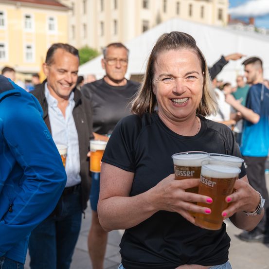 Eine Frau hält drei Biergläser in der Hand, während sie lächelt, umgeben von anderen bei einer Veranstaltung im Freien. Gebäude und Zelte sind im Hintergrund zu sehen.