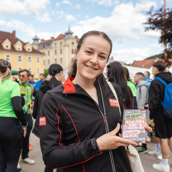 Eine Frau in einer schwarzen Jacke hält eine Zeitschrift in der Hand, lächelt und steht in einer Menschenmenge, vermutlich bei einer Veranstaltung im Freien. Im Hintergrund sind Gebäude und Menschen zu sehen.
