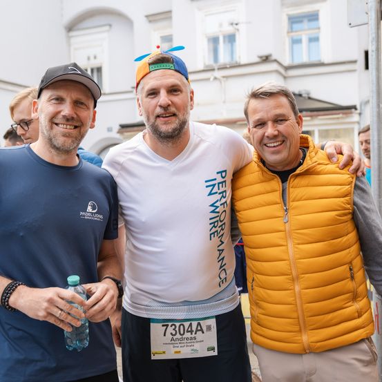 Drei Männer posieren für ein Foto, einer hält eine Wasserflasche. Der Mann in der Mitte trägt ein weißes T-Shirt mit der Aufschrift 'Performance', und der Mann rechts trägt eine gelbe Weste. Dahinter steht ein Gebäude mit Glasfenstern.