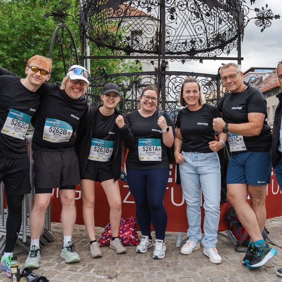Sechs Menschen in Laufshirts mit Nummern posieren vor einer Metallstruktur für ein Foto, wobei ein Mann mit Brille und Turnschuhen lächelt.