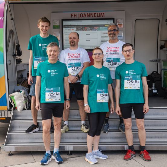 Fünf Personen stehen auf einer Treppe vor einem Gebäude mit der Aufschrift 'FH JOANNEUM'. Sie tragen grüne T-Shirts mit Nummern und 'Invent with Purpose'. Zwei tragen eine Brille und zwei haben eine Uhr. Eine Frau hat eine Tasche auf dem Boden.