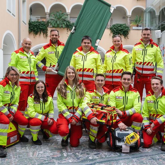 Eine Gruppe von Notfallsanitätern posiert für ein Foto, alle in gelben und roten Uniformen mit reflektierenden Streifen und medizinischen Taschen. Sie lächeln und stehen neben einem grünen Banner.