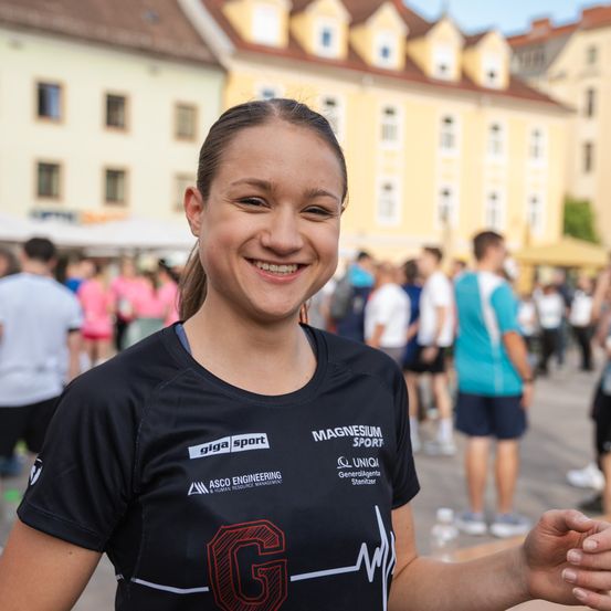 Ein lächelndes Mädchen in einem schwarzen T-Shirt mit dem Buchstaben G darauf steht vor einer Menge. Sie nimmt an einer Veranstaltung teil, mit Gebäuden und Menschen im Hintergrund.