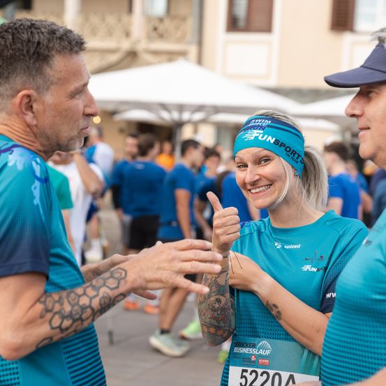 Eine Frau mit Stirnband gibt zwei Männern während eines Laufevents einen Daumen hoch. Sie tragen passende blaue Shirts und stehen inmitten einer Menge. Im Hintergrund sind Gebäude und Schirme zu sehen.