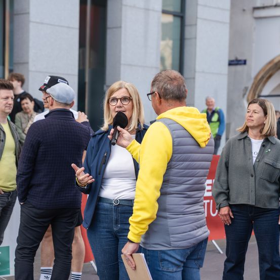 Eine Frau spricht in ein Mikrofon, das ein Mann hält, umgeben von Menschen in einem öffentlichen Raum. Dahinter steht ein Gebäude mit Glasfenstern und einem roten Banner.