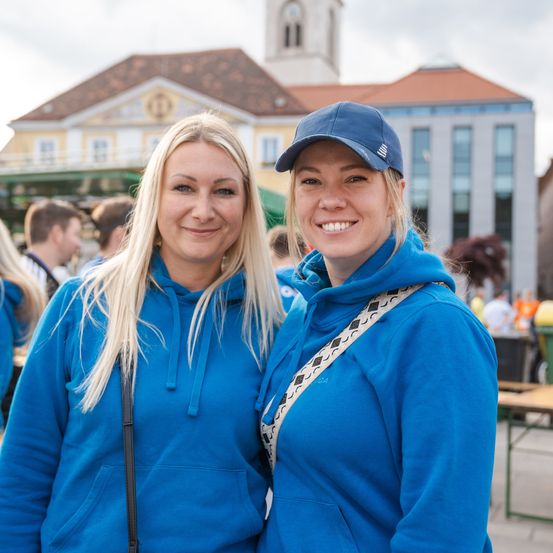 Zwei Frauen in blauen Hoodies lächeln für die Kamera, vor einem Gebäude mit einem Giebeldach und einem Turm stehend. Sie sind in der Nähe eines Mülleimers und Tische mit Menschen im Hintergrund.