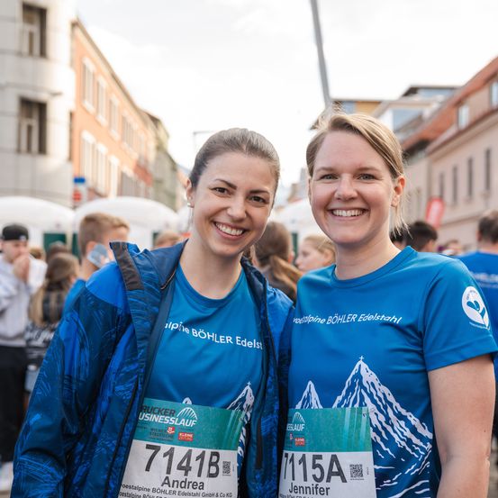 Zwei Frauen in passenden blauen Shirts posieren für ein Foto bei einem Rennevent, mit verwaschenen Menschen im Hintergrund. Beide tragen Startnummern, eine hat eine Jacke an.